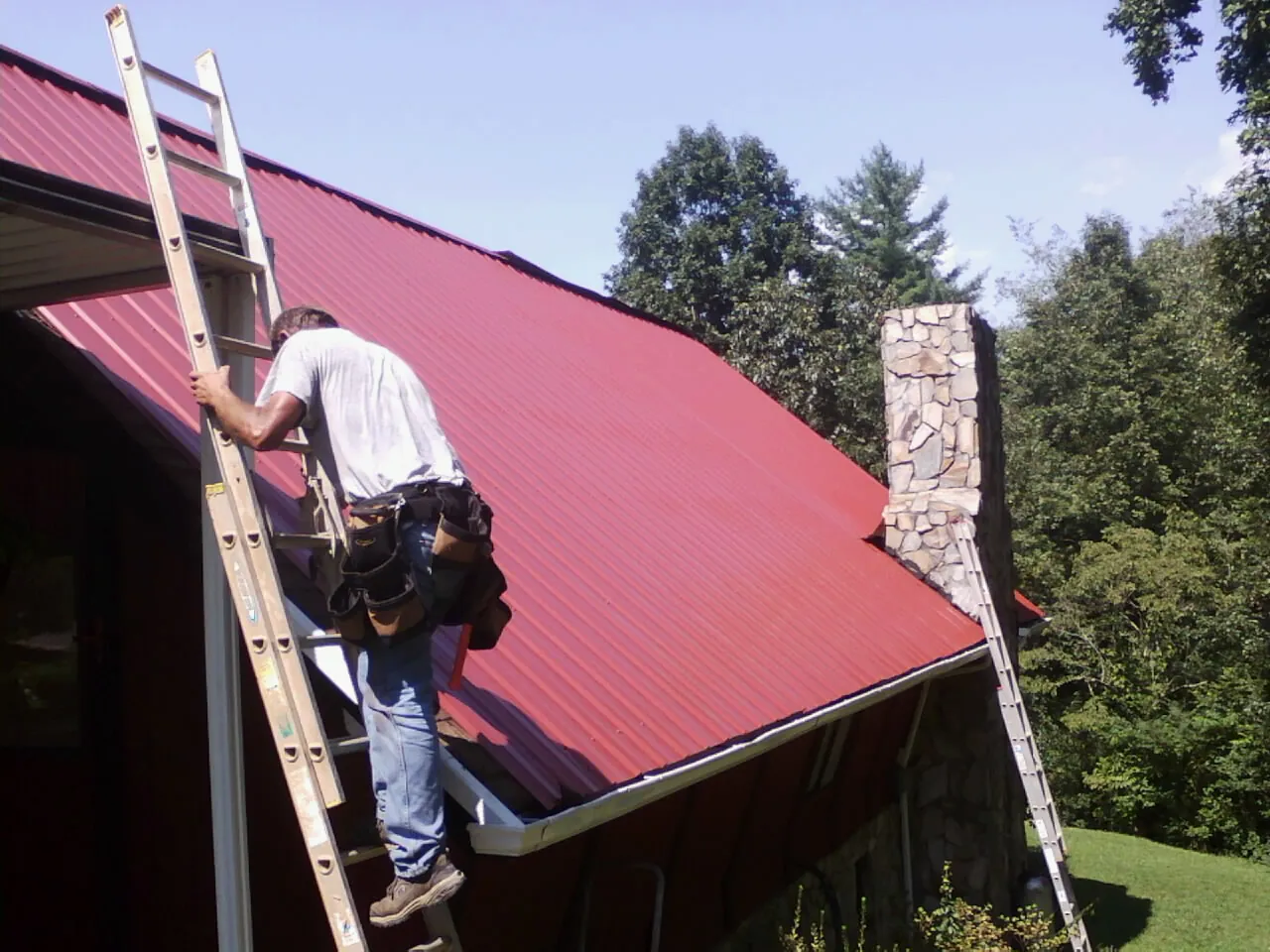 Roofer on a ladder beside a red metal roof and stone chimney — Soffit & Fascia Repair by Roofing Brothers in Cedar